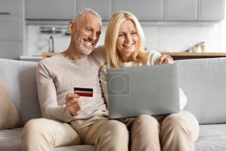 Mature couple happily using a credit card to shop online, sitting on a sofa at home
