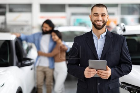 A smiling salesman in the foreground with a tablet, and indian couple interacting with a car in the background