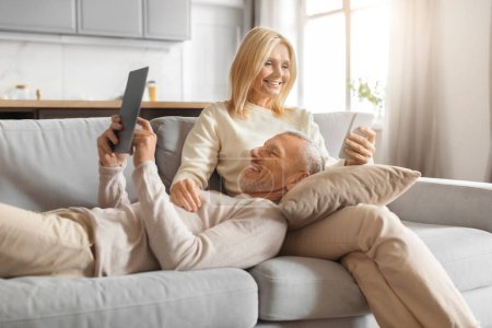 Senior man laughs while browsing a tablet as his partner enjoys her drink, both sharing a fun moment