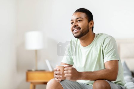 Foto de Alegre joven negro con una camiseta sentada en su cama, sonriendo y mirando hacia otro lado de la cámara - Imagen libre de derechos