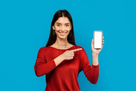 Photo for Woman in Red Shirt Holding Smart Phone And Pointing At Blank Screen, Happy Female Demonstrating App Mockup, Standing On Blue Background - Royalty Free Image