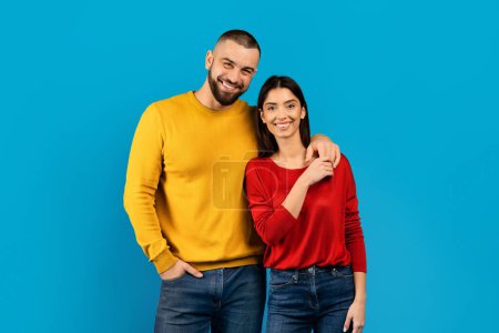 Photo for Smiling couple dressed in colorful sweaters standing confidently on blue background, romantic man and woman posing in studio together against bright backdrop, looking at camera, copy space - Royalty Free Image