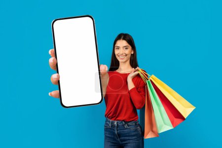 Photo for Woman standing with shopping bags in one hand and blank smartphone in the other, smiling young female multitasking while making purchases, recommending new app, mockup - Royalty Free Image