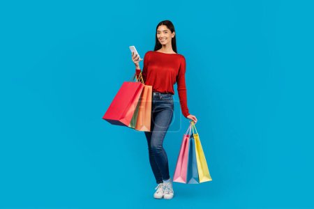 Foto de Mujer sonriente de pie sobre fondo azul, sosteniendo bolsas de compras en una mano y teléfono celular en la otra, espacio para copiar - Imagen libre de derechos