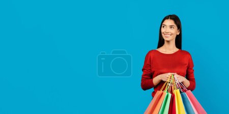 Téléchargez les photos : Femme en haut rouge avec des sacs à provisions colorés regardant l'espace de copie, jeune femme heureuse portant des achats debout isolé sur fond bleu studio, Panorama - en image libre de droit