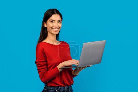 Photo for Smiling young woman wearing red shirt holding laptop in her hands, standing on blue studio background - Royalty Free Image