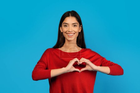 Photo for A woman shaping heart with her hands, expressing love and affection in a simple gesture, happy young female standing isolated on blue studio background, copy space - Royalty Free Image