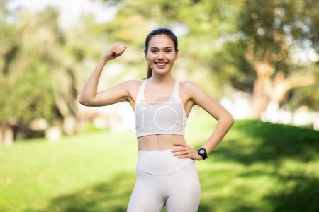 Photo for Glad proud caucasian young woman in a white sports bra and leggings flexes her arm, showcasing her fitness, with a smartwatch on her wrist against a lush park background, outside - Royalty Free Image