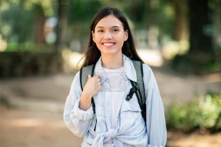 Smiling young european woman with long dark hair wearing a white shirt and light blue jacket tied around her waist carrying a backpack while walking outdoors in a park setting