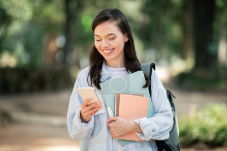 Téléchargez les photos : Engagé jeune fille heureuse européenne jolie étudiante avec une expression joyeuse, en utilisant un smartphone et tenant des carnets pastel, debout à l'extérieur dans un parc verdoyant luxuriant, à l'extérieur - en image libre de droit