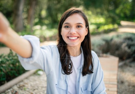 Photo for Radiant glad european young woman with a charming smile extends her arm for a selfie, capturing a moment of joy in a sunlit park, surrounded by rich natural greenery, outside - Royalty Free Image