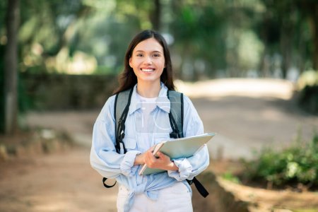 Photo for Content smiling young european lady student with a radiant smile, dressed in white, walking on a path in a park, holding a tablet computer while wearing a backpack, outside - Royalty Free Image