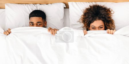 Foto de Divertida pareja afroamericana milenaria escondida bajo una manta blanca en la cama, vista superior - Imagen libre de derechos