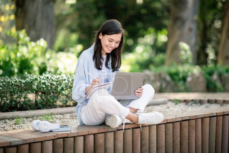 Téléchargez les photos : Une jeune femme européenne concentrée assise sur un banc en bois dans un parc luxuriant, profondément absorbée par des notes écrites sur un tampon tout en utilisant un ordinateur portable, avec un casque et une tasse de café à côté d'elle - en image libre de droit