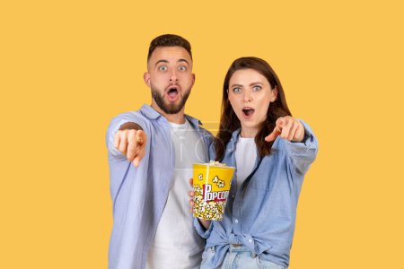 Hombre y mujer mostrando expresiones de conmoción y sorpresa, sosteniendo una caja de palomitas de maíz sobre fondo amarillo