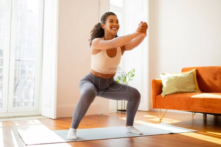 Photo for Active young lady athlete performs squats and stretches her arms forward, showcasing her dedication to fitness and wellbeing in modern domestic setting, exercising in light living room - Royalty Free Image