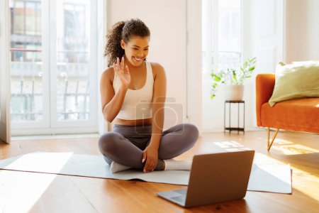 Téléchargez les photos : Femme brésilienne sportive dans les vagues de vêtements de sport bonjour à l'ordinateur portable tout en pratiquant une session de yoga en ligne à la maison, mélangeant la technologie moderne avec le bien-être actif et la routine d'entraînement - en image libre de droit
