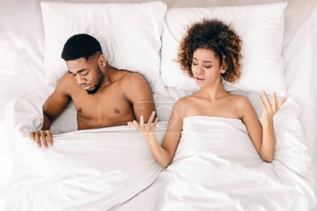 Photo for Sex problem. Upset black lovers lying on bed, woman unsatisfied, sad man looking under blanket, top view - Royalty Free Image