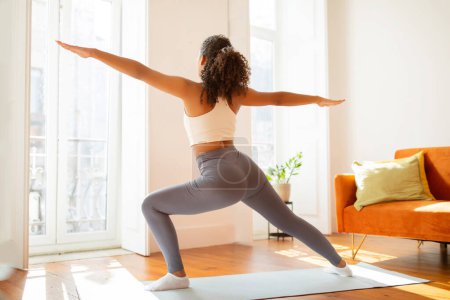 Rear view of woman in fitwear practices yoga doing warrior pose in her living room, focusing on physical and mental wellness with serene zen poses, promoting healthy lifestyle