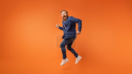 Téléchargez les photos : Homme barbu aux cheveux roux excité portant tenue décontractée sautant et criant joyeusement sur fond de studio orange, exprimant une ambiance énergique positive, s'amusant. Pleine longueur, panorama avec espace de copie - en image libre de droit