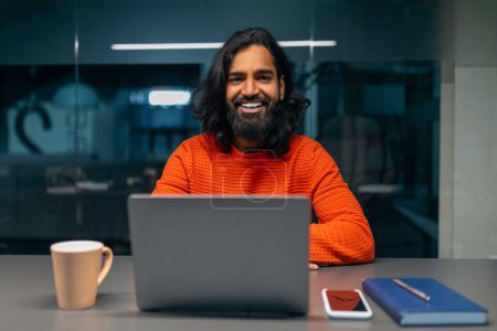 Téléchargez les photos : Homme d'affaires confiant avec un sourire joyeux et un ordinateur portable dans un cadre de bureau décontracté - en image libre de droit