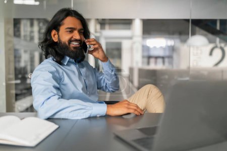 Alegre profesional participar en una conversación por teléfono mientras trabaja con un ordenador portátil en su escritorio de la oficina