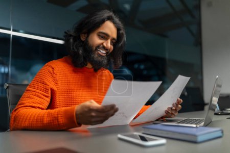 Foto de Hombre de negocios feliz en un suéter brillante leyendo papeles al lado de un ordenador portátil - Imagen libre de derechos