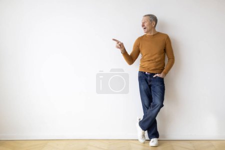 Foto de Un hombre vestido casualmente se levanta contra una pared en blanco, señalando algo fuera de cámara con una mirada de interés o énfasis - Imagen libre de derechos