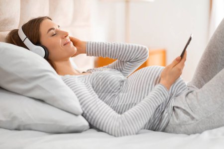 A young adult woman comfortably lying on her bed, using smartphone and headphones smartphone in a well-lit room, capturing a relaxed everyday moment