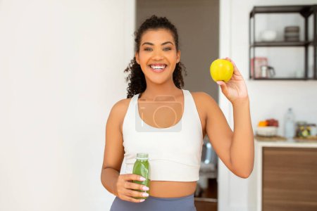 Photo for Happy sporty young lady embraces wellbeing and healthy nutrition, holding a fresh smoothie and apple fruit, showcasing her healthy diet and lifestyle in modern kitchen after morning workout - Royalty Free Image
