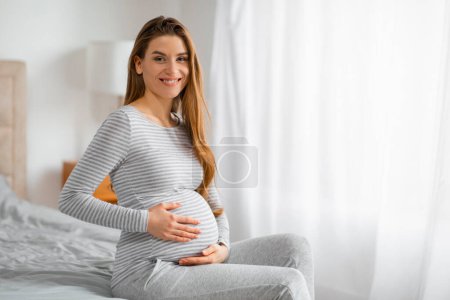 Schwangere sitzt bequem auf dem Bett, die Hände auf dem Bauch und trägt ein angenehmes Lächeln