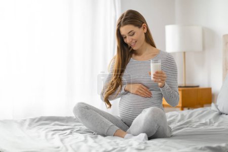 Photo for A content pregnant woman in comfy attire holds a glass of milk in a bedroom, conveying a sense of health and prenatal nutrition - Royalty Free Image