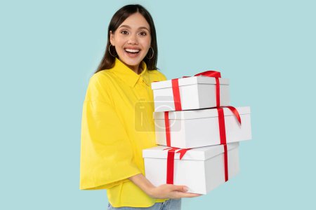 Photo for Happy trendy young brunette woman in chic yellow shirt, holding stylish white gifts with bold red ribbons, posing against blue backdrop. Celebration and holidays concept. - Royalty Free Image
