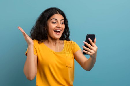 Delighted woman holds her smartphone, seemingly video calling with an excited expression on teal