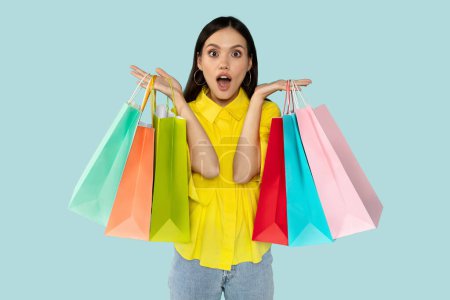 Überraschte hübsche brünette junge Frau in gelbem Hemd mit bunten Einkaufstaschen und Grimassen, die den Black Friday Sale genießt, blauer Hintergrund. Konsum, Einzelhandel