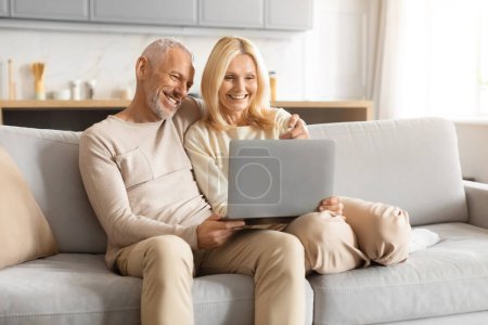 Photo for Joyful mature couple having fun while sitting and interacting with a laptop computer on the couch - Royalty Free Image