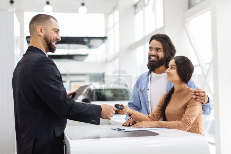 A cheerful young eastern couple is handed car keys by a dealer at a modern car showroom, signifying a new purchase