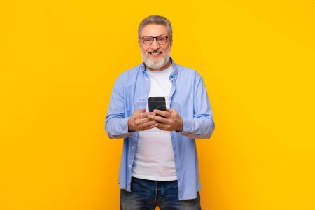 Téléchargez les photos : Homme âgé aux cheveux gris sourit tout en naviguant sur le téléphone debout dans un cadre studio jaune, souriant à la caméra tout en appréciant la communication mobile moderne, textos sur le gadget de téléphone portable - en image libre de droit