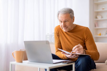Photo for An elderly male in casual wear appears focused as he works on a laptop while holding a notebook in a well-lit room - Royalty Free Image
