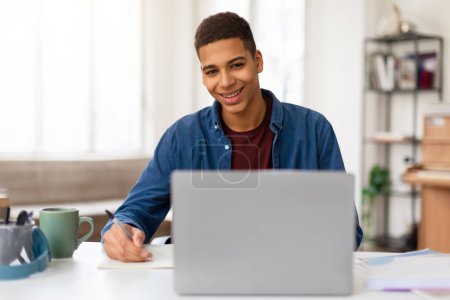 Fröhlicher junger Mann macht sich Notizen in einem Notizbuch, während er in einem hell erleuchteten Raum an seinem Laptop arbeitet