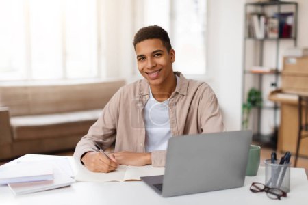 Foto de Alegre estudiante negro masculino en atuendo casual escribe notas en el cuaderno mientras trabaja en el ordenador portátil, exudando sensación de productividad y satisfacción - Imagen libre de derechos