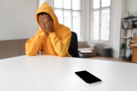 Photo for A person in distress sits with their head in hands, a smartphone lying on a table in the foreground, signaling a difficult moment - Royalty Free Image