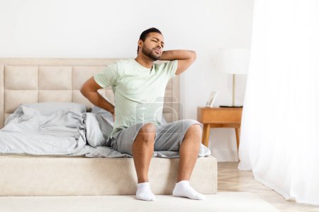 Téléchargez les photos : L'homme afro-américain étire le dos alors qu'il est assis sur le lit, souffre après s'être réveillé, dans un simple cadre de chambre - en image libre de droit