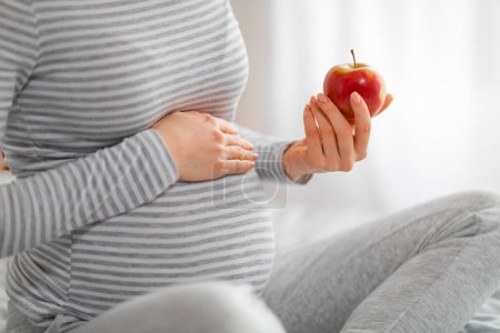Foto de La imagen muestra a una mujer embarazada serena sosteniendo suavemente una manzana roja, lo que significa elecciones nutricionales inteligentes - Imagen libre de derechos