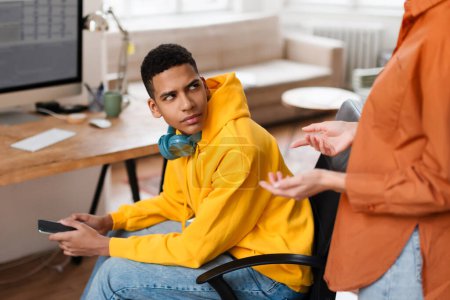 Téléchargez les photos : Adolescent sceptique gars à capuche jaune écoute maman tout en tenant smartphone, se tournant pour s'adresser à un conférencier invisible lors de la discussion à la maison - en image libre de droit