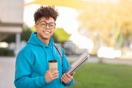Téléchargez les photos : Un gentil jeune étudiant brésilien sourit chaleureusement tout en tenant du café et des cartables d'école lors d'une promenade en ville - en image libre de droit