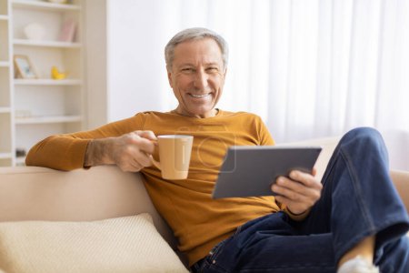 Téléchargez les photos : Un aîné souriant tient une tasse à café tout en utilisant une tablette, exsudant un sentiment de détente et de plaisir dans sa maison - en image libre de droit