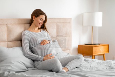 Téléchargez les photos : Une femme enceinte sereine s'assoit confortablement sur un lit, tenant son ventre dans une chambre bien éclairée, suggérant un moment de repos et de liaison avec son enfant à naître - en image libre de droit