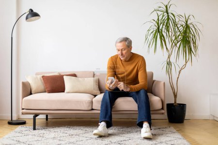 Foto de Un hombre mayor con un atuendo casual se sienta en un sofá con un teléfono, luciendo comprometido y cómodo en un entorno hogareño - Imagen libre de derechos
