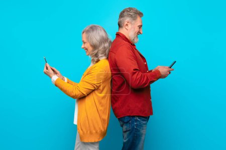 Téléchargez les photos : Homme et femme âgés défilant et bavardant sur leurs téléphones portables, représentant la joie des médias sociaux et la connectivité dans la vie des personnes âgées, debout dos à dos avec des smartphones sur fond de studio bleu - en image libre de droit